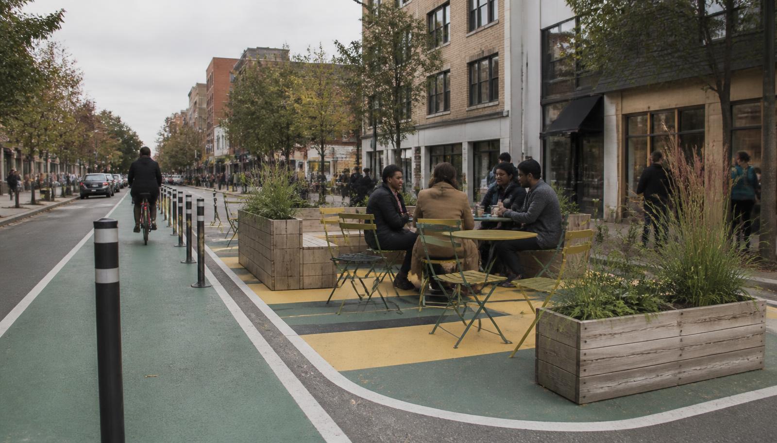 A temporary parklet of timber planters and moveable chairs occupying two parking bays beside a pop-up cycle lane.